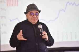 Salvador Di Stefano dando una charla con micrófono en mano, usando sombrero y anteojos, con gráficos económicos proyectados de fondo.