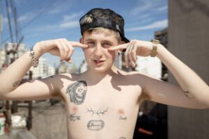 El rapero Dillom en una terraza sin remera mostrando sus tatuajes en el torso, con gorra hacia atras y manos en su rostro