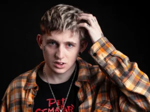 Dillom con camisa a cuadros marrón y collar, expresión seria que refleja su estética ligada al trap y rap argentino
